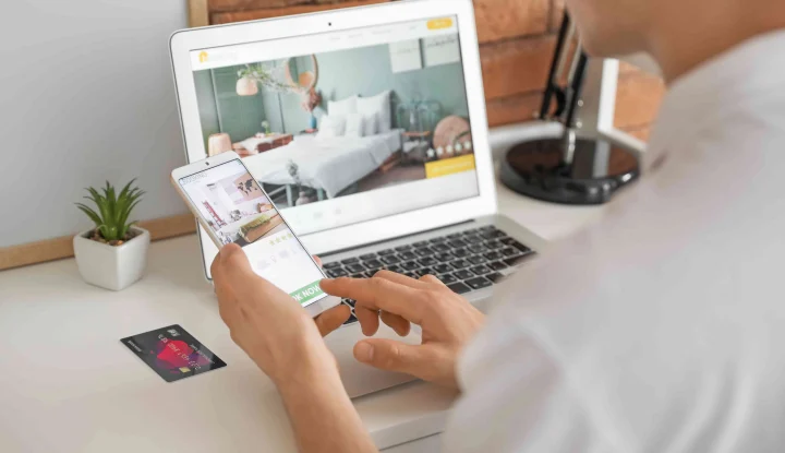Boost your hotel's holiday revenue: 8 tips to increase sales this season. A person is looking at a hotel website on their phone with their computer and credit card next to them.