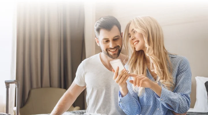 Why Hospitality Solutions has the Best Hotel Management Software - A man and a woman sit on a hotel bed smiling at their phone - Hospitality Solutions
