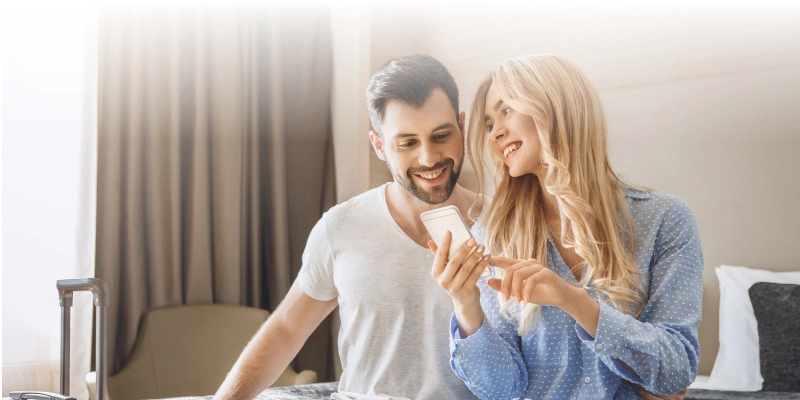 Why Hospitality Solutions has the Best Hotel Management Software - A man and a woman sit on a hotel bed smiling at their phone - Hospitality Solutions